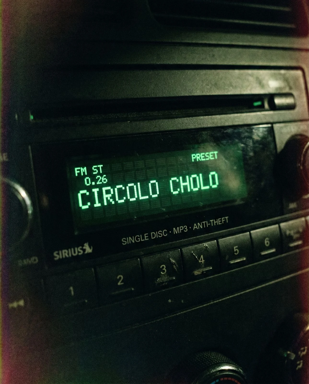 Un'autoradio nella penombra: sul display verde la scritta «CIRCOLO CHOLO» in caratteri a punti, sopra la fila dei tasti numerati da 1 a 6.