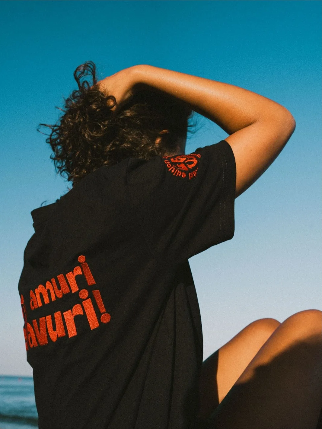 Una maglietta nera vista da dietro, sulla spiaggia: la frase «amuri peffavuri!» in rosso sulla schiena, un braccio alzato e i capelli che coprono il volto.