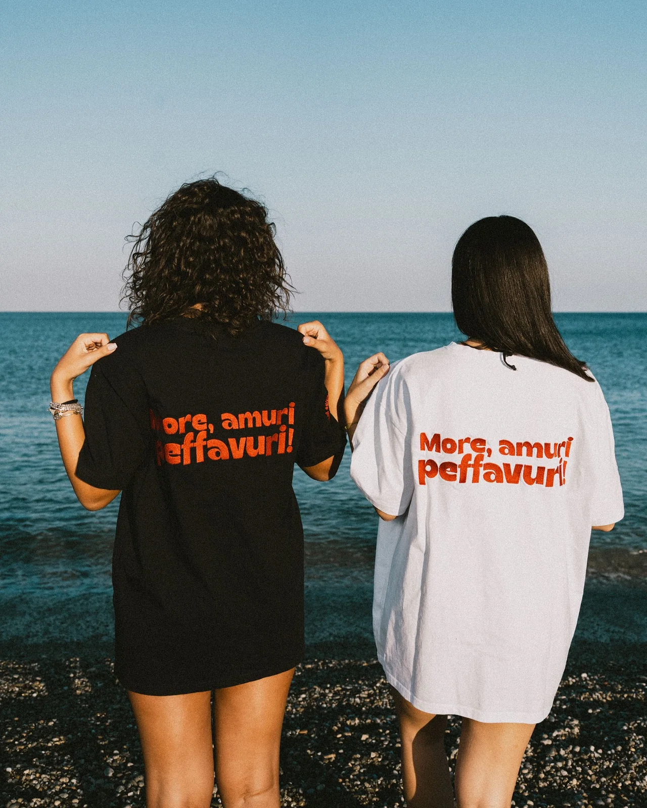 Due magliette oversize, nera e bianca, viste da dietro sulla spiaggia: sulla schiena la frase «More, amuri peffavuri!» in rosso.