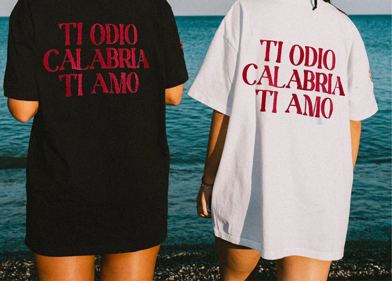 Le schiene di due magliette, nera e bianca, davanti al mare: la frase «Ti odio Calabria ti amo» in rosso su tre righe.