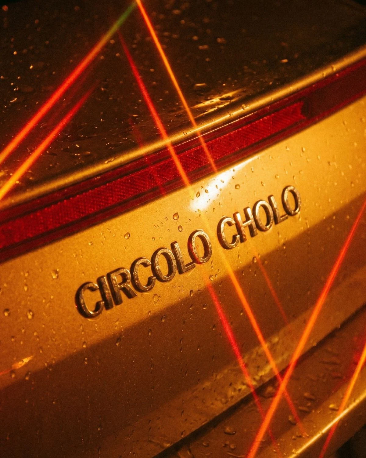 La scritta «CIRCOLO CHOLO» in lettere cromate sul retro di un'auto bagnata, attraversata da strisce di luce rossa e arancione.