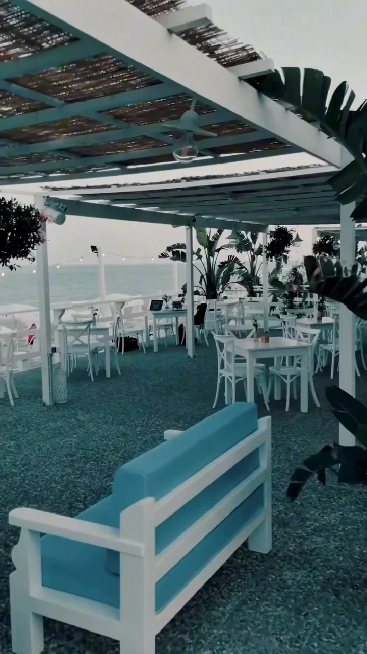 La terrazza sotto il pergolato: in primo piano una panca bianca con cuscini azzurri, dietro tavoli e sedie bianchi tra le piante e, sullo sfondo, il mare con una fila di luci appese.