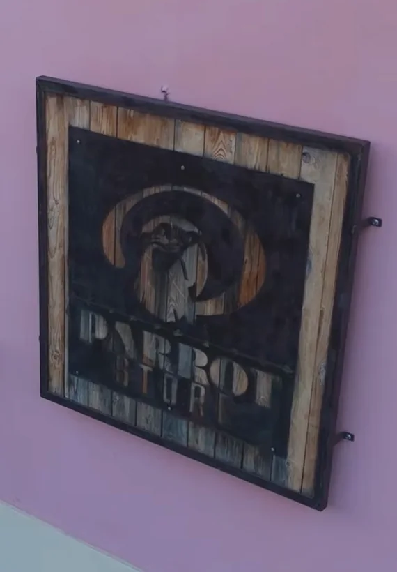 L'insegna del negozio sulla facciata rosa: una cornice di assi di legno e una lastra scura in cui sono ritagliati il pappagallo e la scritta PARROT STORE.